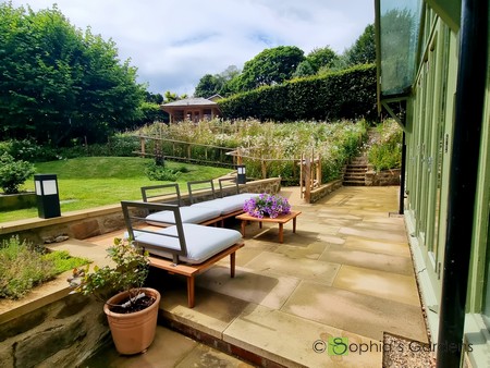Before image shows a vast expanse of green grass, over a multi leval garden. There are two trees within the grassed areas, a low stone wall to the front of the image with steps to the front left. A blue clipboard with white paper and a pen lay on the grass at the front of the image. The after image shows a smooth stone wall to the left of the image, leading to a gravel pathway that zigzags up the sloped garden to a natural wood summerhouse with pointed roof, glass doors and windows. A wooden fence accompanies the pathway and lush green grass features on either side of the path. Click image for more details.