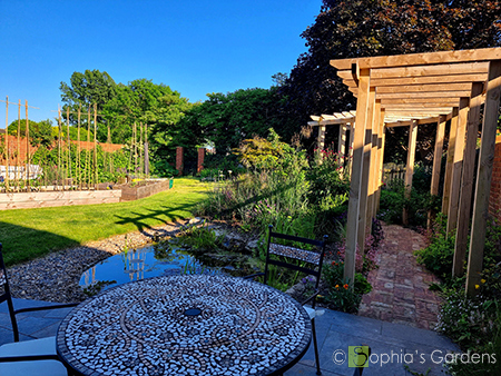 An overgrown garden with an old greenhouse to the left top corner and netball net at the bottom right. plain, simple garden layout with large trees on the boundary. After image shows bright blue skies with a small mosaic patio table at the bottom with wooden pergola above the pathway to the right leading to the top of the garden. Raised kitchen garden beds to the left with a green lawn in the centre. Click image for more details.